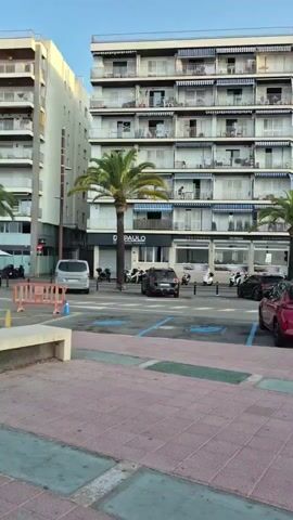 The video begins with a view of a street scene featuring several multi-story buildings, palm trees, and parked cars. In the foreground, a red car is parked in a designated spot. Further back, several other vehicles are visible, including a black van and a white SUV. A person in a white shirt and dark pants walks away from the camera. The buildings appear to be hotels or apartment complexes, with balconies and windows. The sky is clear and blue, indicating daytime. The video pans slightly to the right, showing more of the street and buildings.