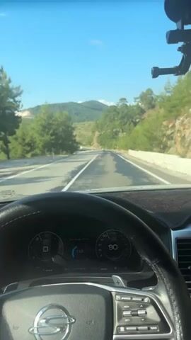 A first-person perspective video shows a driver's hands on the steering wheel of a car as it travels down a paved road. The dashboard displays the gear as D, and the speed is 91 km/h. The road is flanked by trees and hills under a clear blue sky. A white van is visible in the distance on the road ahead. The camera is mounted above the dashboard, providing a clear view of the road and the surrounding landscape.