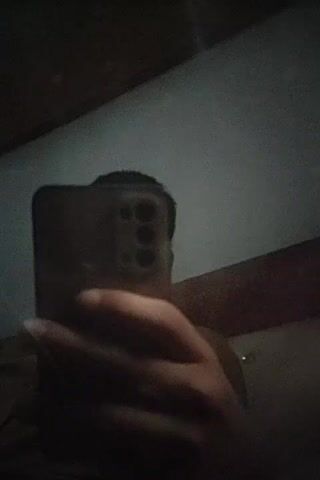 The video shows a person holding a smartphone in front of a mirror, taking a selfie. The person's face is partially obscured by the phone, but their head and a portion of their arm are visible. The background appears to be an indoor setting with a gray wall and a dark ceiling. The lighting is dim, creating shadows and a sense of low visibility. The smartphone has a distinctive camera bump on the back, featuring multiple lenses arranged in a rectangular pattern. The person's hand is clearly visible, gripping the phone. The camera angle suggests the selfie is being taken from a slightly low perspective. The video is very short, consisting of only a few seconds of footage.