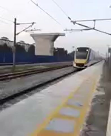 The video shows a train arriving at a station. The train is white with red and blue stripes. It is a modern electric train with large windows. The station platform is made of concrete and has yellow markings. There are overhead power lines for the electric train. The weather appears to be overcast. The train is moving from right to left in the frame and slows down as it approaches the platform. The camera is stationary and positioned on the platform.