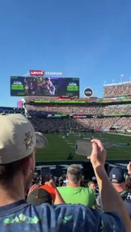 The video depicts a crowd of people in a packed stadium, likely attending a football game. Most people are holding up their phones, possibly recording or taking photos of the field. The stadium is brightly lit, and a large screen displays images. Some people are wearing sports jerseys. The overall environment suggests an exciting and energetic event.