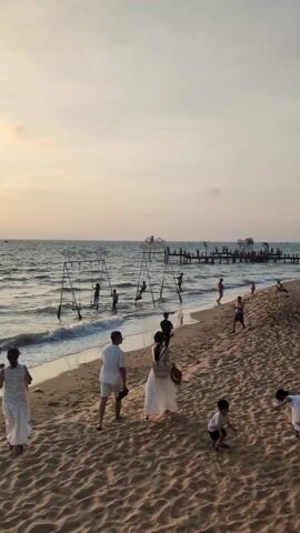 The scene captures a beach at what appears to be sunset or twilight, with a sandy shore meeting the sea. Several people are present. Some are walking along the shoreline, while others are engaging in activities near or in the water. Notably, there are swing sets placed in the shallow water where children are swinging.  A wooden pier extends into the sea in the distance.