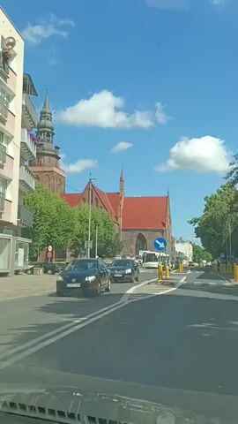 The video shows a street scene with a brick church with a steeple in the background. A residential building with balconies is on the left. Cars are driving on the road in front of the church. A person can be seen on a balcony on the residential building. There are trees and street signs visible. The sky is blue with some clouds.