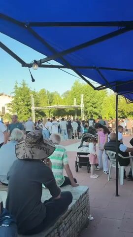 The video shows a crowd of people watching a folk dance performance on an outdoor stage. The stage is a metal structure with a canopy roof. The dancers are wearing traditional costumes. Some audience members are standing while some are seated on a stone ledge. There are trees and bushes visible behind the stage. The weather appears to be sunny.