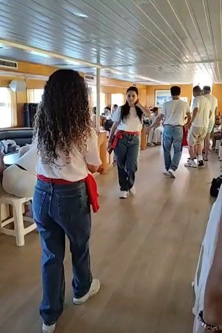 The video shows the interior of a boat with a wooden floor and a white ceiling. There are people of various ages, most of them dressed casually in jeans and t-shirts. Some people are seated on benches along the sides of the boat, while others are standing. In the foreground, two young women are facing each other. The woman on the left has long, curly brown hair and is wearing a white t-shirt, blue jeans, and white sneakers. She has a red sash tied around her waist. The woman on the right has dark, straight hair and is wearing a similar outfit: a white t-shirt, blue jeans, and red sash. The woman on the right is in motion, appearing to be dancing or performing. In the background, several men are also dressed in jeans and white t-shirts. The atmosphere appears to be relaxed and celebratory. The lighting suggests it is daytime, with light coming in from windows on the side of the boat.