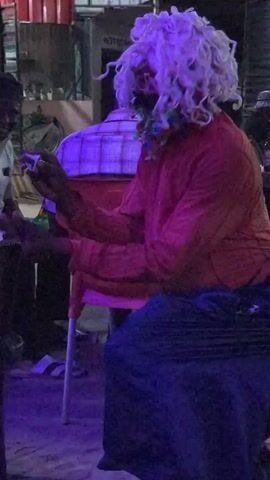 The video shows three people in what appears to be a bar or restaurant. One man is wearing a white shirt and is holding a green bottle. Another man is sitting in a red chair, wearing a plaid shirt. The third person is wearing a red shirt, a long dark skirt, and a light-colored wig. Their face is obscured by something colorful, possibly makeup or a mask. The music is playing in the background.
