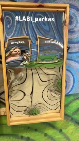 This is a short video showing a woman taking a selfie in a funhouse mirror. The mirror distorts her reflection, making her appear stretched and warped. The background shows a wall with a blue and white swirling pattern and a wooden floor. There are other distorted reflections of the woman in smaller mirrors in the background. The hashtag #LABI_parkas is visible at the top of the mirror. The woman is wearing a grey t-shirt and denim shorts, and is holding a phone in her right hand. Her left hand is on her hip. She is wearing white sneakers.