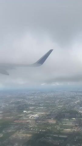 The video shows a view from an airplane window. The wing of the plane is visible through a dense layer of clouds. Below the clouds, a cityscape is visible, appearing hazy and somewhat obscured.