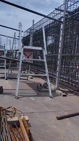 The video shows a construction site with a large steel rebar cage. In the foreground, there is a rolling ladder and some bundles of rebar. A construction worker wearing a hard hat and safety harness walks behind the rebar cage, carrying a white plastic bag. In the background, there are electrical towers and some fencing. The weather appears to be cloudy.