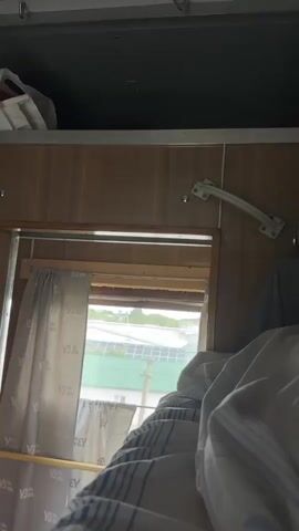 The video shows an interior view from a train compartment looking out a window. The bed with blankets is in the foreground, and a curtain hangs to the left of the window. Outside, buildings and trees are visible, along with a roof covered in solar panels. The compartment appears to be made of wood paneling, with storage space above the window.
