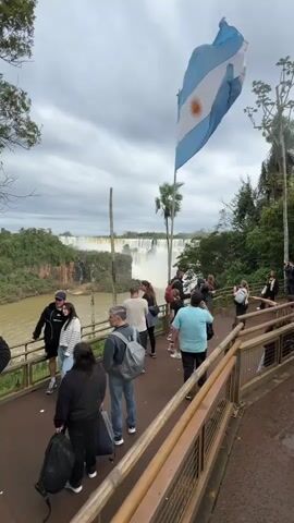 A group of tourists stand on a boardwalk, some taking pictures. In the background, a waterfall can be seen. An Argentinian flag flies in the foreground.