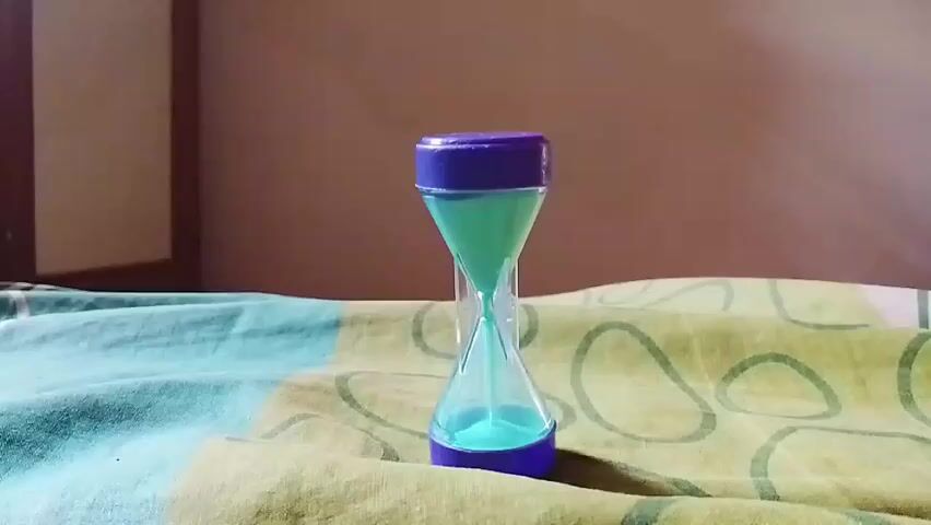 This is a close-up, static shot of a small, hourglass timer. The hourglass has purple caps on the top and bottom, and a clear glass middle section containing teal-colored sand. The sand is currently flowing from the top bulb to the bottom bulb. The hourglass is placed on a patterned surface, likely a blanket or bedspread, with a background that appears to be a wall. The lighting is natural, suggesting it is daytime. There are no people or other objects visible in the frame. The video appears to be a short time-lapse or slow-motion recording of the hourglass in action.