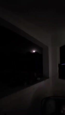 The video shows a night sky with a bright moon visible through the trees. It is dark outside, and the moon is shining brightly. The trees are silhouetted against the moon, and the sky is dark. There are some objects on a windowsill in the foreground.