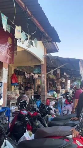 The video shows a busy outdoor market scene. In the foreground, several motorcycles are parked, their seats visible. A person wearing a maroon shirt and a helmet is walking away from the camera towards a stall. Behind the motorcycles, market stalls are set up with awnings. One stall has a large red banner with text and an image. Various goods are displayed on shelves and tables in the stalls, including clothing and what appear to be food items. Several people are present in the market, some appearing to be vendors and others shoppers. The environment is an open-air market with structures providing shade. The time of day appears to be daytime, with sunlight visible.