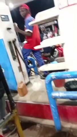 The video shows the exterior of a gas station at night. A young man wearing a red shirt with "OMNILIFE" printed on the back, blue pants, and a red cap is standing on a blue motorcycle. He is leaning on a gas pump and appears to be operating it. A small brown jug sits on the ground next to the pump. Another person wearing a red shirt is briefly visible in the foreground, partially obscuring the view. There are additional people and vehicles in the background, near another pump. The gas station has white pillars and a red-and-white sign. There is a blue metal barrier in the foreground, with a bucket on it.