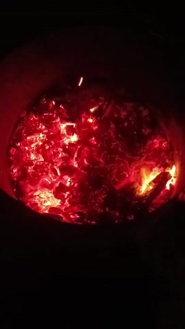 A close-up shot focuses on a pile of glowing red embers within a dark, cylindrical container. The embers are hot and appear to be wood or charcoal. The edges of the container are visible as dark red, suggesting it is also heated. The lighting is dim, with the intense glow of the embers providing the primary illumination. There are no people or distinct objects visible besides the burning embers and the container. The scene is static, with no discernible movement or action beyond the subtle flickering of the flames within the embers.