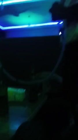 The video shows a dimly lit room with colorful LED lighting, likely a lounge or hookah bar. There is a table in the center covered with drinks and hookahs. Two people are present, one wearing a white hat and the other dressed in white, both are sitting on a dark couch. The man in the white hat picks up what appears to be a lighter and looks at his phone. Music plays in the background.
