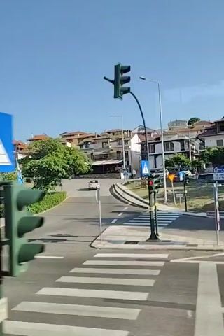 The video shows a view from a moving vehicle on a sunny day. The scene transitions from a road intersection with traffic lights and pedestrian crossings to a road alongside a hill with buildings and trees. A silver car drives past on the road. The video ends with a view of trees and a white banner attached to a fence.
