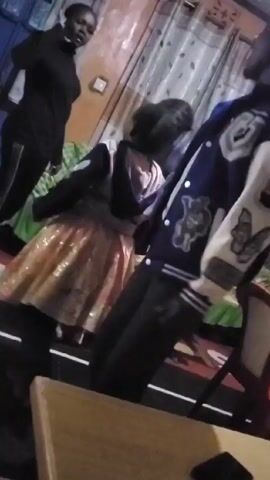 The video shows a blurry indoor scene with several people present. An adult woman wearing a black turtleneck and dark pants stands to the left. A child in a dress with a sparkly orange skirt is in the center, facing away from the camera. To the right is a person wearing a blue jacket with white sleeves and patches, including one with a butterfly design. The people appear to be moving or dancing to music. The room has patterned curtains, green bed covers and a wooden chair is visible in the foreground. The lighting is dim.