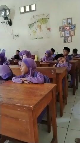 The video shows a classroom scene filled with young children, likely elementary school students, predominantly dressed in purple uniforms, some wearing hijabs. They are seated at individual wooden desks and chairs, facing forward, suggesting they are engaged in a lesson or activity with a teacher who is not visible in the frame.

The children appear attentive; some have their mouths open as if speaking or reciting, while others are looking around or smiling. Water bottles are visible on several desks. The classroom walls are light-colored and decorated with simple paintings, including a large tree with the word "Baca" (Indonesian for "Read") painted underneath, and some flowers. A world map and framed pictures are also displayed on the wall. A wall-mounted fan is visible in the upper left corner, and natural light enters through windows, indicating it is daytime. The overall atmosphere appears to be one of active learning and engagement within a school in Citerep, Indonesia.