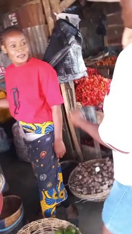 The video shows an open-air market. Several people are browsing and selecting produce. There are baskets of green avocados, red tomatoes, and other items on display. A woman with long braids, wearing a white t-shirt and blue shorts, examines something in her hand. Other individuals are visible in the background, and the environment appears to be bustling with activity. The lighting suggests it is daytime.