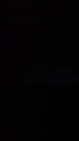 The video shows a dimly lit landscape at night. In the distance, there is a faint glow, suggesting the presence of lights from a settlement or buildings. The sky is dark, with some clouds visible, and there appears to be a silhouette of trees or structures on the horizon. The foreground is mostly black, indicating a dark, possibly rural, environment. There are no people or discernible objects in the foreground. The overall scene is very dark and lacks clear detail.