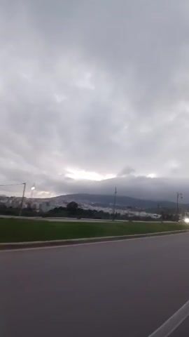 A first person view from a moving vehicle shows an asphalt road and a grassy median with various plants and street lights. Behind the median is a cityscape at the base of a mountain or large hill. The sky is very cloudy and gray. Two cars pass by, one green and one black.