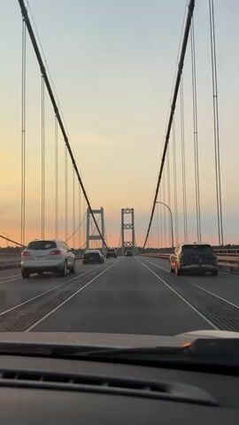 This is a POV video from a car driving across a suspension bridge during sunset. The sky is a gradient of orange and yellow with scattered clouds. The bridge has two large towers in the distance and is lined with multiple cars driving in the same direction. The camera shows the dashboard and a stuffed animal in the foreground.