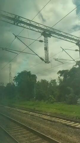 The video shows a view from a moving train. The train tracks are visible in the foreground. On the right side of the tracks, there is a stretch of green grass and trees. In the background, a tall communication tower can be seen. The sky is overcast with clouds. The video pans slightly to the left as the train moves.