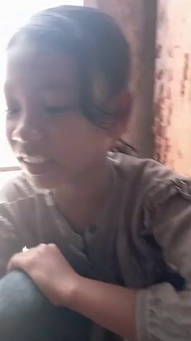 A young girl with dark hair pulled back in a ponytail is sitting on the floor near a white wall or window. She is wearing a gray, long-sleeved shirt. She looks to the side, smiles, then looks forward and appears to make a rude gesture with her hand. The background is somewhat blurred, but there appears to be a wooden wall or structure nearby.