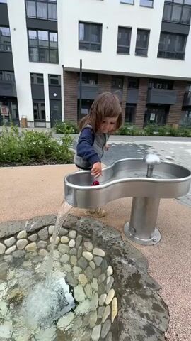A young child with long brown hair and wearing a grey vest, a blue long-sleeved shirt, and leopard-print pants plays with a water fountain. The child is standing on a sandy-colored surface next to a modern building with white and brown sections and large windows. The fountain has a metal basin with a curving spout where water is pouring into a circular, rock-lined pool below. The child is reaching into the spout with their right hand, and a small red object is visible in their hand. The water in the pool is splashing, creating a small fountain effect. The child appears to be focused on playing with the water.
