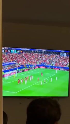The video shows a large flat-screen television displaying a soccer match. The game is between Portugal (POR) and Spain (ESP), with the score currently 0-0. The scene on the TV shows players in white and red uniforms on a green soccer field. A crowd of spectators wearing red is visible in the stands. The bottom of the screen shows a player with curly, dyed hair in profile. The TV is mounted on a wall in a dimly lit room. The bottom edge of the frame shows the heads and shoulders of two people watching the television.
