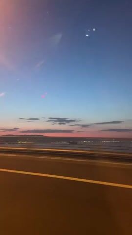 From a moving vehicle, the video captures a twilight sky displaying a gradient of blue to orange hues above a body of water speckled with lights. A distant coastline is visible. The vehicle travels on a road marked by yellow lines. A safety rail runs along the road's edge, closest to the camera. The scene suggests a bridge or elevated road at dusk.