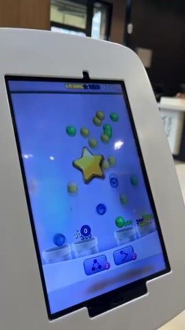The video shows a close-up of a white tablet device mounted on a stand. The tablet screen displays a game with colorful bubbles and a yellow star. The background is out of focus but appears to be an indoor setting, possibly a restaurant or cafe. In the far background, a counter with the word "CASSA" is visible, suggesting a point of sale or service area. The lighting is bright, with a ceiling light prominently featured in the upper portion of the frame. The tablet is the primary focus, and the game on its screen is dynamic, with bubbles moving and changing position.