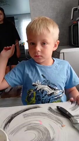 A young boy with blond hair, wearing a blue t-shirt and black shorts, is sitting at a table. A woman wearing a black t-shirt and black shorts stands behind him, with her left arm extended. The boy turns his head to his right, and the woman places her right hand on his shoulder. The boy then turns back towards the camera. The camera is positioned at table level, looking towards the boy and the woman. The environment appears to be a kitchen or dining area, with a patterned tablecloth on the table, a bowl with some food, and kitchen appliances in the background.