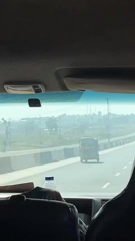 The video is filmed from inside a vehicle, looking out through the windshield onto a multi-lane highway in Sabon Gora, Nigeria, during the daytime. The sky appears hazy, suggesting dry or dusty conditions.

In the foreground, parts of the vehicle's interior are visible, including the dashboard, a water bottle, a rearview mirror bracket, and a sun visor. The camera is positioned behind a passenger seat, with a person's shoulder or the back of a head briefly seen in the lower right corner.

The highway ahead is a paved road with clear white lane markings, stretching into the distance. Concrete barriers separate the directions of traffic on the left side of the road. The road appears to ascend gently.

Initially, a yellow auto-rickshaw (tuk-tuk) is visible directly in front of the vehicle, moving in the same direction. As the video progresses, the auto-rickshaw is no longer immediately ahead, and other vehicles, including another auto-rickshaw, can be seen further down the long, straight highway.

The landscape surrounding the road consists of scattered trees, sparse vegetation, and some distant structures, all viewed under the hazy conditions. Power lines and utility poles are also visible along the horizon. The scene captures travel on a main road through a semi-rural environment. No legible text is visible on any objects or signs.