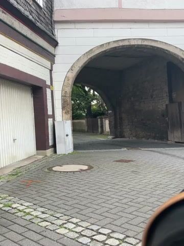 This video pans across a European street scene. The cobblestone pathway leads through an arched stone gateway into a narrower lane. The buildings on either side are a mix of styles, with some featuring timber framing and others modern facades with stucco and window shutters. A white station wagon is parked on the right side of the street.

Notable details include:

*   **Scene:** A quiet street, likely in a historic European town.
*   **Setting:** The architecture suggests Germany, specifically the region of Lower Saxony. The presence of cobblestone streets, archways, and timber-framed buildings are characteristic.
*   **Time of day/Weather:** The lighting suggests it's daytime, possibly overcast or early morning, as there are no strong shadows.
*   **Objects:** A white car, various building features (archways, shutters, timber framing), a street sign, manhole covers, and a garage door.
*   **Text:** A blue and white street sign is visible. The text on the sign reads: "Zufahrt zum Grundstück bis 3,5t frei" and "Bedingung zum Parkplatz". This translates to "Access to the property free up to 3.5 tons" and "Condition for the parking lot." The car also has a license plate with "W" and "KH" visible, likely indicating its registration area. A sign on one of the buildings says "PLAN HOUSE."
*   **Activity:** The video is a slow pan, suggesting exploration or documentation of the location. There are no people actively engaged in activities shown in the frame.
*   **Emotions:** The overall mood is peaceful and tranquil, given the quiet street and the absence of bustling activity.