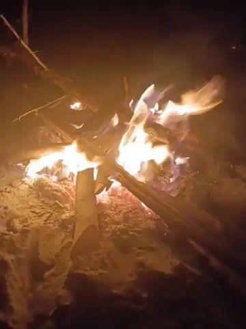 The video shows a campfire burning on what appears to be sand at night. The flames are bright, and there are several pieces of wood arranged in a teepee shape. Some small sparks are visible flying upward.