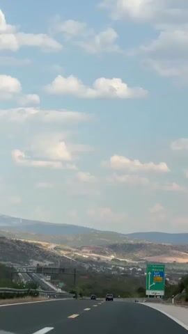 A vertical video captures a car's perspective on a highway. The road curves gently to the right, flanked by guardrails and sparse vegetation. In the distance, rolling hills and mountains form a hazy backdrop under a bright, cloud-dotted sky. A green highway sign with white lettering and arrows is visible on the right side of the road, indicating destinations and route numbers. Two cars are seen ahead on the highway, traveling in the same direction. The scene suggests a daytime journey through a rural or semi-rural landscape.