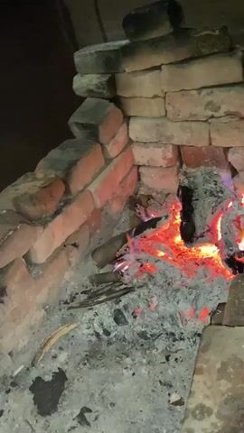 The video captures a close-up view of a brick fireplace with a fire burning inside. The fireplace is constructed from red bricks, some of which appear weathered or discolored. The fire is composed of glowing red embers and small pieces of wood, with a visible orange and yellow flame in the center. Ash covers the bottom of the fireplace and the surrounding hearth. The lighting is dim, with the fire providing the primary light source. The camera is positioned slightly above and to the side of the fireplace, looking downwards. There are no people or animals visible in the video. The scene appears to be indoors, given the brick construction and likely the presence of a hearth.