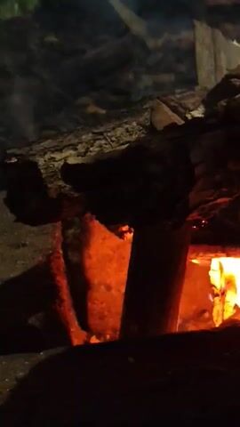 The video shows a close-up of a burning wood fire. Flames are visible, along with glowing embers and charred logs. Smoke drifts upwards. The scene appears to be outdoors or in an open area where burning is permitted. There are no people or other identifiable objects in the frame.