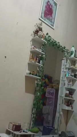 A video captures a bedroom with a vanity mirror, shelves, and decor. The mirror is tall and framed with white lights and artificial ivy. Two corner shelves are mounted on the wall to the left of the mirror, holding various bottles and containers. The right side of the mirror also has shelves attached, filled with similar items. A large wardrobe or cabinet stands to the right of the mirror, with shelves displaying books and other objects. A framed picture of two people, possibly a couple or family, hangs on the wall above the left shelves. A small table with boxes and items is visible in the foreground to the left. Several trash cans are placed near the wardrobe. The room appears to be decorated in a personal style, with an emphasis on organization and display of personal items.