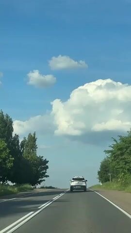 A white car drives down a two-lane road in the countryside on a sunny day. Green trees and fields line the road. The sky is bright blue with large white clouds.
