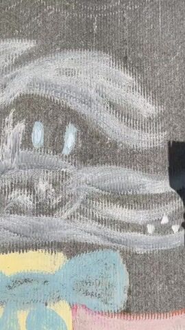 A hand is shown painting a chalk drawing on a textured gray surface, possibly a sidewalk. The drawing appears to be of a face with large blue eyes and a white, fluffy outline. The hand holds a brush with a light-colored handle and dark bristles. The brush is being used to spread a gray substance, possibly water or paint, over a section of the drawing. The lighting suggests it is daytime, with a shadow cast by the person painting. Other chalk colors, yellow and blue, are visible at the bottom of the frame.