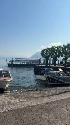 The video captures a serene daytime scene along a sunny waterfront, likely a large lake, under a clear blue sky.

In the initial shot (0:00), a concrete or paved shoreline is visible in the foreground. To the left, a white cabin cruiser-style boat with a brown trim and dark windows is moored close to the camera. On the right, a larger, two-deck passenger ferry, predominantly grey and white, is either docked at a pier or slowly moving. Several green trees line the pier area, and part of another small boat is visible further right.

As the camera pans (0:01-0:02), two nearly identical white cabin cruiser-style boats come into view, moored side-by-side directly at the concrete shore. They are connected by a short wooden gangway. Both boats feature cabin structures and open deck areas. A small Italian flag is visible on the stern of the boat on the left.

The final segment of the video (0:02-0:03) shows a different white cabin cruiser, with a red lifebuoy prominently attached to its side, moored alongside a small pier structure on the left. Part of another boat is visible on the right.

Throughout the video, a wide expanse of calm, blue water stretches across the middle ground. In the distance, majestic mountains rise, their lower slopes dotted with greenery and what appear to be buildings. The clear weather and bright sunlight suggest a pleasant day. The scene evokes the peaceful atmosphere typical of a scenic Italian lake region, consistent with the provided location context of Binda, Italy. No visible text is present in the video.