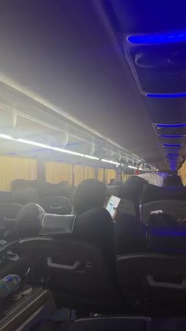 The video begins inside a bus, showing rows of empty seats and yellow curtains. Blue lights are visible along the ceiling. The camera then pans to the right, showing a blurry view of the bus interior. Next, the camera shows a nighttime view from inside the bus, looking out at a well-lit restaurant with the sign "DECOR OF DELHI RESTAURANT" prominently displayed. Several cars are parked in front of the restaurant, and a few people are visible. The camera continues to pan, showing more of the restaurant exterior and the surrounding area, which appears to be a parking lot with some debris on the ground.