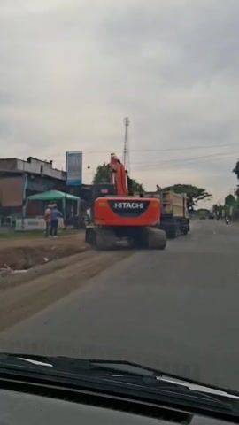 The video, recorded from inside a vehicle, shows a construction scene on the side of a road. An orange Hitachi excavator is visible, partially on the dirt shoulder and partially on the asphalt. Next to it, a beige dump truck is parked. A few people are standing near the excavator and dump truck, appearing to be workers. The truck begins to drive forward, moving past the excavator. The road is paved, and there's a dirt area on the right. In the background, there are buildings, trees, and a tall communication tower. The sky is overcast.
