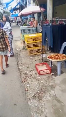 A shaky, low-angle video shows a street market scene. In the foreground, a person in a checkered shirt and shorts walks away from the camera, their legs showing some redness. To the left, a person in a hijab and patterned pants walks towards the camera. In the middle ground, several stacks of yellow and green plastic crates are visible, some containing what appear to be chickens. A person in a grey tank top leans over one of the crates. Beyond the crates, a market stall with a pink umbrella and a rack of blue pants can be seen. The ground is a rough, dirty asphalt surface with debris. In the right foreground, a red plastic tray with some food items is on the ground. In the background, a motorbike is parked, and other market stalls and people are present. The overall impression is a busy, outdoor market.
