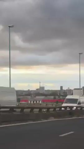 The video shows traffic on a highway, with a city skyline visible in the background beneath a cloudy sky. The camera is positioned to the side of the highway, likely within a vehicle, capturing the movement of cars and trucks. Some tall buildings and a tower can be seen in the distance, blending with the grey and white sky. The highway has a concrete barrier with a red stripe on the other side, and light poles are visible along the side of the road.