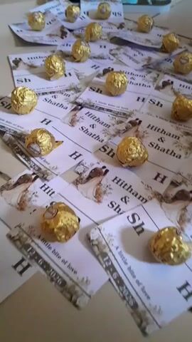 The video shows a collection of Ferrero Rocher chocolates scattered across a surface. Each chocolate is individually wrapped in gold foil. They are placed on small cards or invitations that feature the names "Hitham" and "Shatha" along with the initials "H Sh." The cards also display an image of a bride and groom and a tagline that reads "A little bite of love." In the background, a white bowl filled with white tulips can be seen, along with some decorative white items with ornate patterns. The overall scene appears to be related to a wedding or a special celebration.